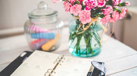 Zdjęcie stołu na którym stoją: wazon z kwiatami, słoik z kredą do pisania oraz leży organizer z kalendarzem.